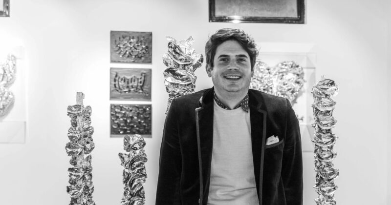 Arthur Dupuy, artiste sétois, en veste de velours devant des sculptures argentées évoquant des coquilles d'huîtres, photographie en noir et blanc